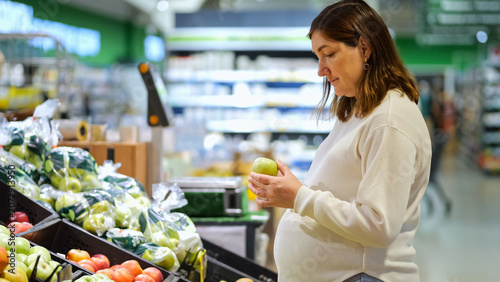 Pregnant woman selecting a green apple from a fruit display in a brightly lit supermarket, focusing on healthy eating and nutrition during pregnancy for a balanced lifestyle