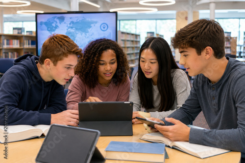 Group of diverse high school students studying together with digital tablets in a library.