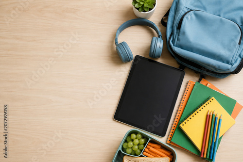 Flat lay of school supplies with tablet headphones backpack and healthy lunch on wooden desk.