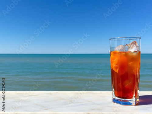 Wide angle shot. A minimalist composition featuring a single tall glass of Aperol Spritz placed on the far right third of a polished white marble ledge. calm turquoise ocean and clear blue sky