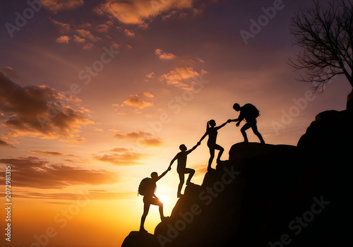 Silhouettes of three hikers helping each other climb up a steep mountain peak during a beautiful golden sunset