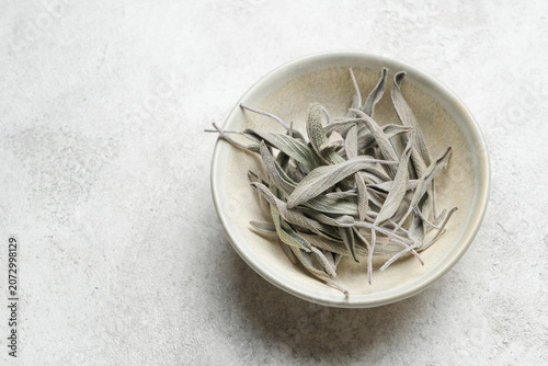 Dried sage herb leaves are neatly placed in a beige bowl on a light gray surface, highlighting their natural color and texture for culinary applications