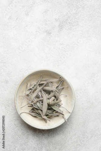 Sage herb leaves are neatly placed in a shallow bowl on a textured surface, highlighting their distinct shape and muted colors