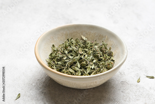 Dried organic thyme leaves displayed in a shallow ceramic bowl on a textured surface, highlighting the green color and delicate structure of the herb