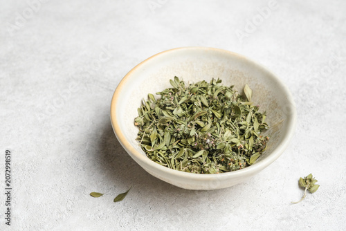 Dried thyme herb leaves are displayed in a round ceramic bowl on a light textured surface, with a few scattered leaves around the bowl