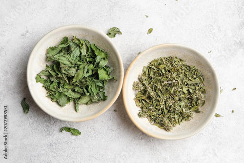 Two ceramic bowls contain dry organic mint herbs, one with vibrant green leaves and the other with dried foliage, placed on a light gray surface