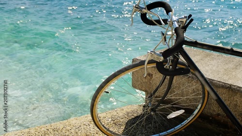 Close-up of vintage road bicycle handlebar and golden-rimmed wheel leaning on a stone pier, sparkling turquoise sea behind.