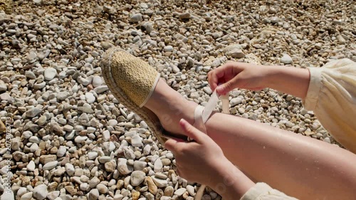 Close-up overhead shot of woman tying woven sandal laces on a foot surrounded by smooth pebbles on the beach, natural light.