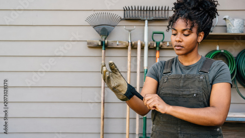 A young woman puts on gardening gloves while standing in front of a wall with organized garden tools and hoses. She appears focused and ready to work outdoors.