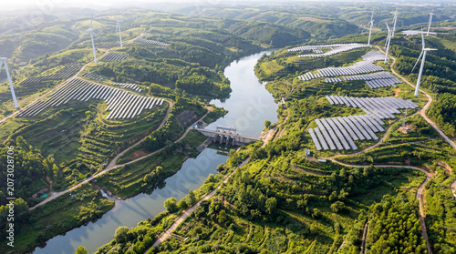 Aerial view of integrated energy systems featuring solar panels and wind turbines alongside a river, surrounded by lush green hills and agricultural land