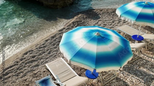 Blue and white beach umbrellas with sun loungers on a sunny pebble beach near clear turquoise sea water, handheld travel footage of a peaceful summer resort coastline.