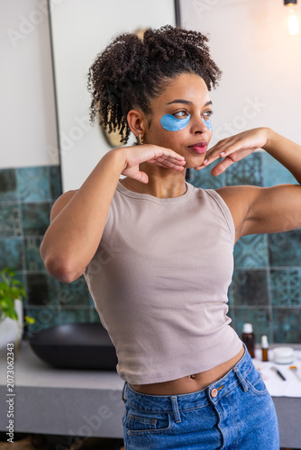 African American woman standing at home vanity wearing tank, posing with blue eye patches
