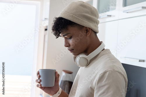 Fotografía Man holding ribbed mug, wearing beanie and headphones, glancing in bright kitche