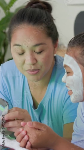 Vertical video: Showing mother guiding LED mask use after handheld prompt in bed, daughter reaching