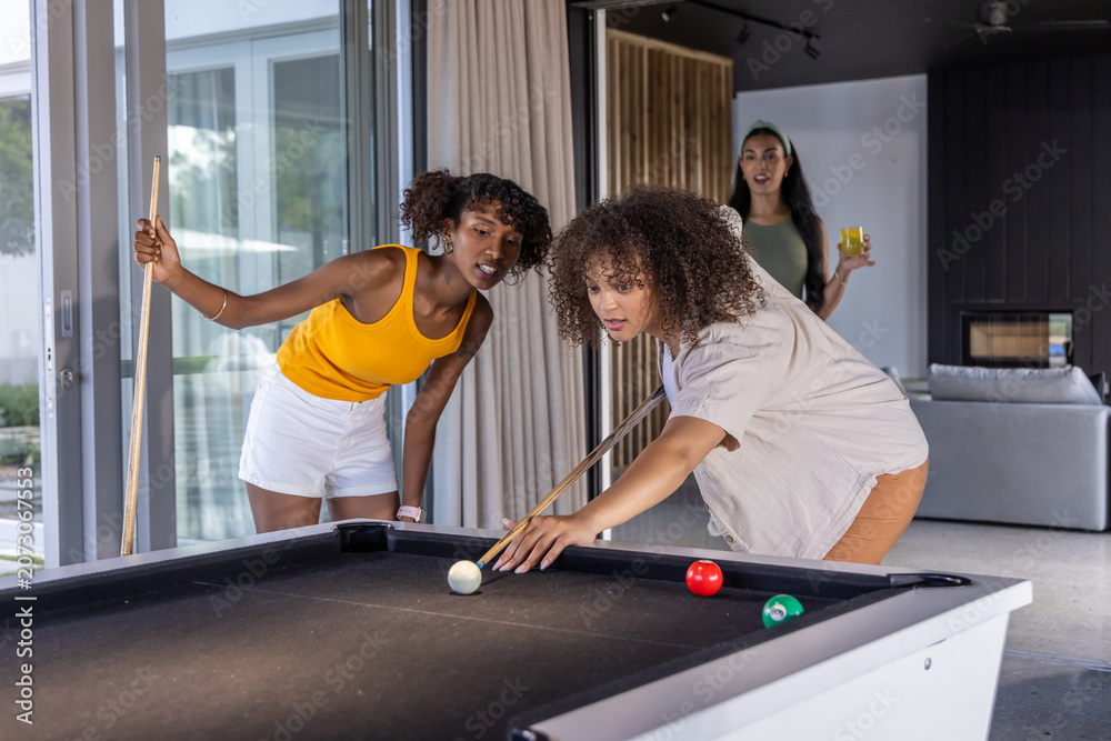 Naklejka premium Diverse female friends playing pool at home on black table with cues, wearing yellow tank top