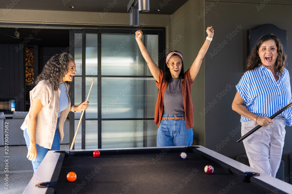 Naklejka premium Three female friends playing in modern game room, celebrating at pool table with cues and balls