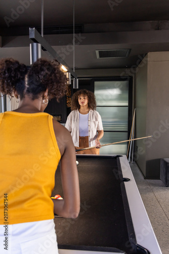 Diverse female friends wearing yellow tank playing pool in game room with cues, copy space