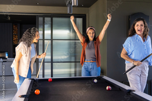 Three female friends playing in modern game room, celebrating at pool table with cues and balls