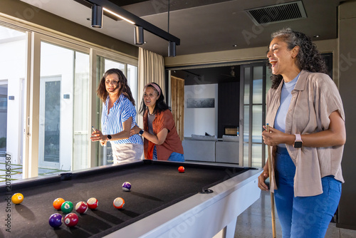 Diverse female friends holding cues and playing pool around black-felt table at home, laughing