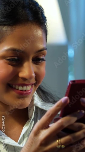 Indian businesswoman checking mobile phone at desk in modern cozy office, Asian female glued to smartphone while multitasking during office hours, smiling with positive body language in workplace