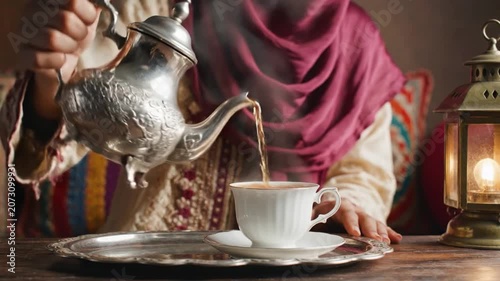 Close-up motion scene shows ornate engraved metal teapot pouring hot tea into white porcelain teacup and matching saucer on decorative tray, with lantern glow, textile backdrop, partial hand visible