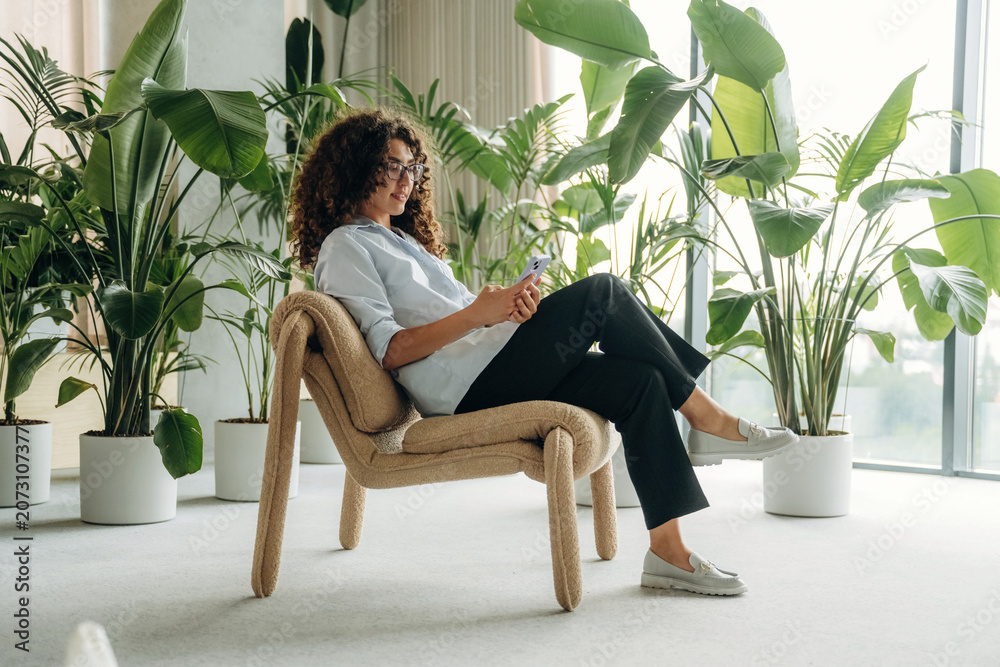 Naklejka premium Young woman with curly hair sitting on a modern chair using smartphone surrounded by large indoor plants in a bright, airy room with natural light