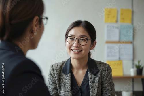 Happy Asian woman with glasses talks with younger woman at desk. Professor advises student, discusses career growth, future job prospects in university office