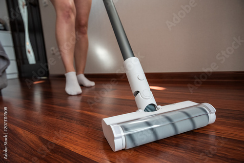 Obraz na plátně Low angle shot of a white motorized floor brush of a cordless vacuum cleaner on a dark wooden floor, woman in white socks cleaning living room for perfect home hygiene and spotless lifestyle