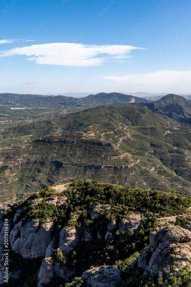 Fototapeta premium Breathtaking aerial vista of the lush green mountain terrain and valleys in the Montserrat region of Catalonia, Spain, under a clear, bright sky with light wispy clouds.