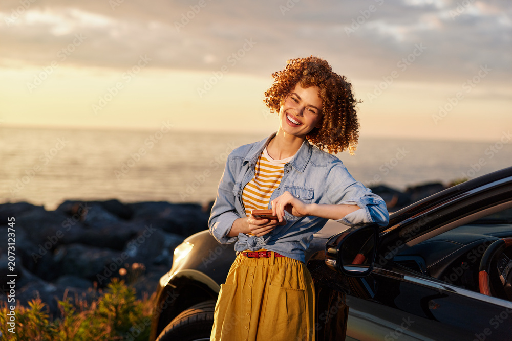 Fototapeta premium Smiling woman with curly hair leaning beside a car at sunset, wearing a denim shirt and mustard skirt. Outdoor lifestyle, travel vibe, carefree mood, warm colors, golden hour lighting.