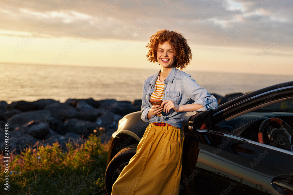 Fototapeta premium Cheerful woman outdoors by a car, smiling in a denim jacket and yellow skirt, sunset light, coastal breeze, candid lifestyle moment, bright natural colors and relaxed posture