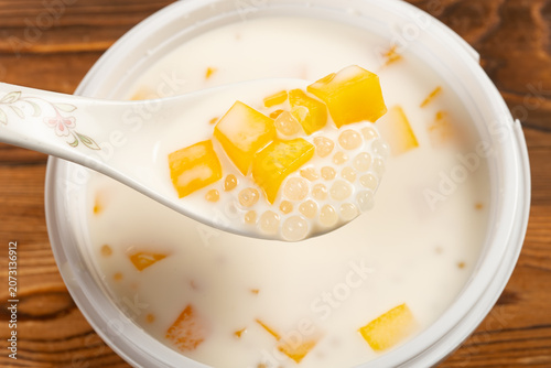 Coconut milk sago dessert with diced mango is held on a floral ceramic spoon above a plastic bowl