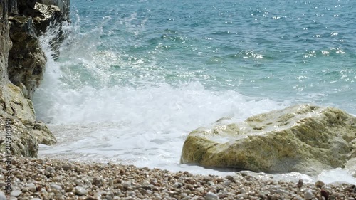 Waves rolling onto a pebble beach between rocky boulders and a cliff, clear turquoise water, handheld nature B-roll shot.