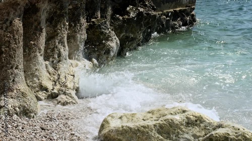 Waves crashing against a rugged limestone cliff with boulders and pebbles in the foreground, handheld close-up, turquoise sea.