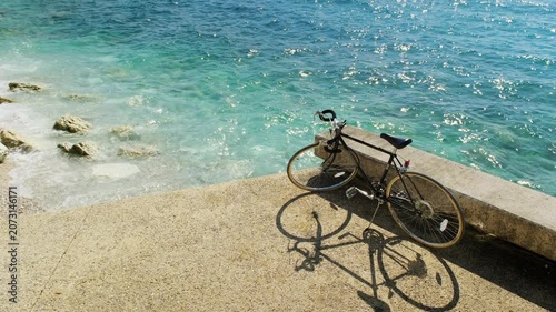 Classic road bicycle with golden rims and handlebar bag leaning on a pier, dramatic shadow on concrete, turquoise sea behind.