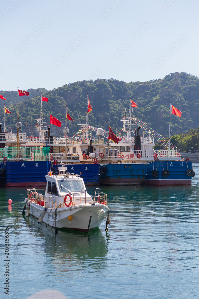 Fototapeta premium Dalian, China-June 5, 2026: Small motorboat moored in calm harbor with fishing vessels and China flags against green hills at Fisherman’s Wharf is one of the city’s most photogenic seaside spots
