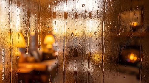 Close up of water drops and condensation trickling down a window pane with a cozy warm blurry living room and lamps in the background