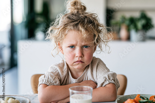 Young girl with curly hair looks upset sitting at a table with a glass of milk and plates of food in a bright kitchen setting.