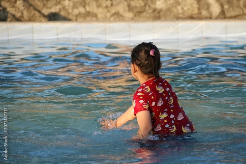 Young girl in a red swimsuit shirt splashes and plays in a clear backyard pool, enjoying warm sunlight and summer fun. Casual candid scene conveying joy, playfulness, childhood and leisure.
