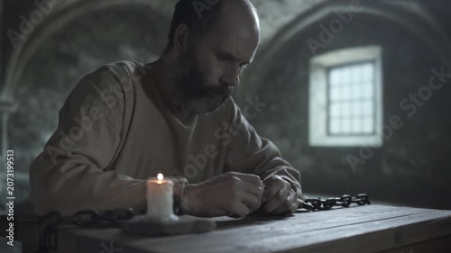 Bearded medieval prisoner in rusty cuffs breaking bread over a rustic wooden table, eating a broken piece while sitting next to a solitary burning candle, inside a gloomy prison room