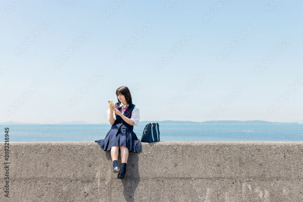 Fototapeta premium 海辺の防波堤でスマートフォンを見る女子高校生