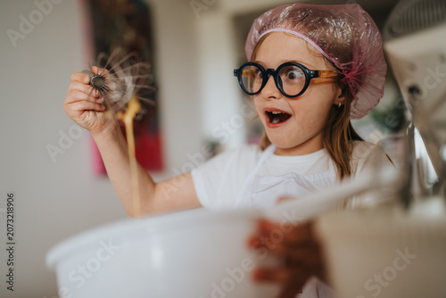 Cuadro en lienzo A surprised young girl wearing glasses and a shower cap holds a whisk over a mixing bowl while baking at home