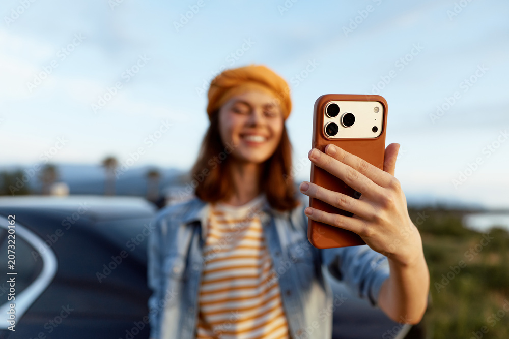 Fototapeta premium A cheerful woman takes a selfie outdoors, smiling at her phone in bright daylight. A casual denim jacket and striped shirt convey a relaxed, everyday lifestyle moment as the road and distant