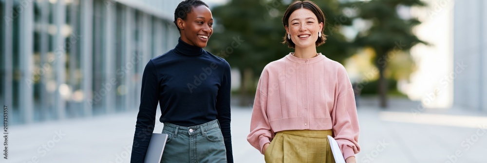 Naklejka premium Two professional women walking side by side outside with laptop and papers for workplace banner and team background use