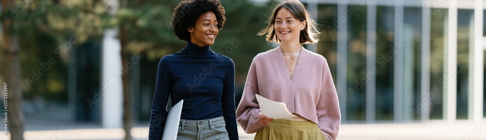 Fototapeta premium Two women walking outdoors smiling while holding papers for workplace lifestyle background and education banner design