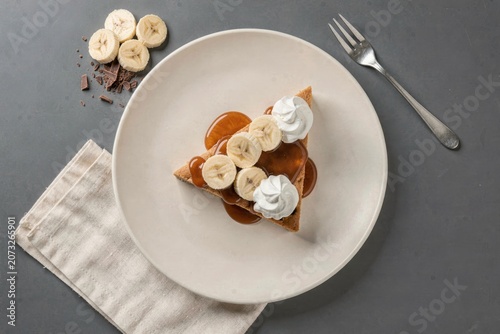 Plate with Banoffee Pie topped with sliced bananas, whipped cream, caramel sauce, fork next to plate. Concept of banana caramel cream pie with biscuit, showcasing dessert creativity.