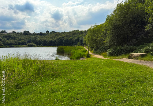 A serene path winds along the edge of a calm lake, embraced by vibrant greenery. Soft, fluffy clouds float lazily across a partly sunny sky in Horton Bank Country Park, Bradford, UK.