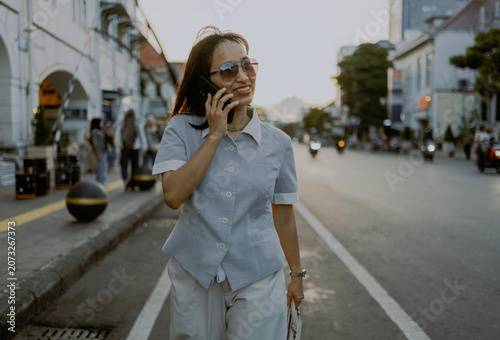 Fashionable style adult Asian Businesswoman commuting in sunglasses and casual button down shirt talking by phone call while walking in city at sunsets.