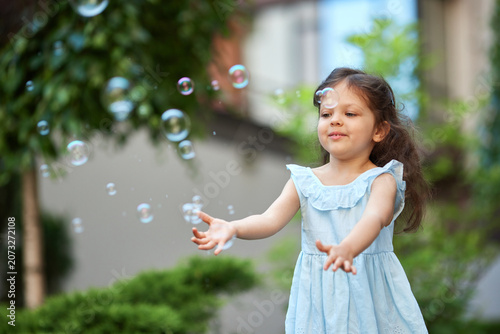 Young girl in a light blue dress with long hair joyfully blowing soap bubbles in a sunny green garden, carefree summer playtime