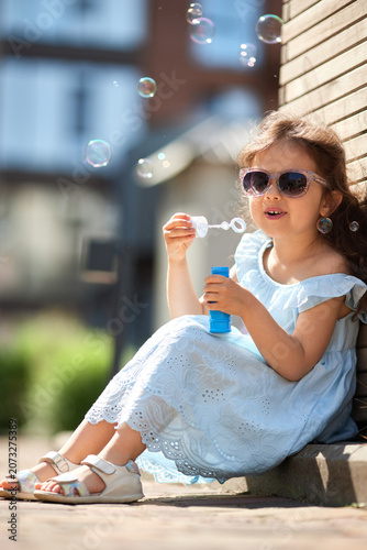 Little girl in blue dress and sunglasses sitting outdoors on a sunny day, joyfully blowing bubbles and smiling with carefree delight